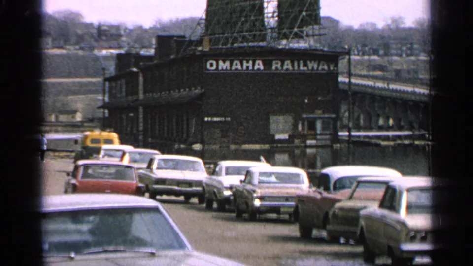 1965 Minnesota home-movie view of traffic moving past the Omaha Railway building, with bridgework and window framing visible.