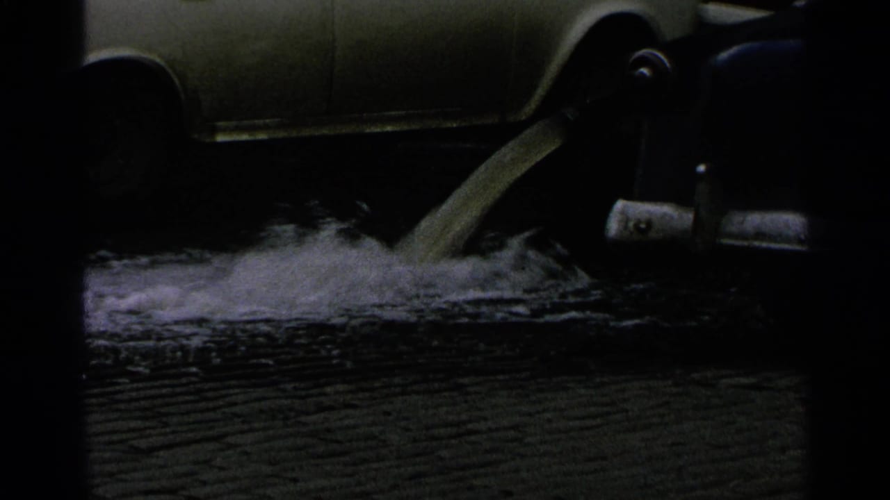 Foamy liquid pours onto wet pavement near the side of a parked car in 1965 Minnesota 8mm footage.