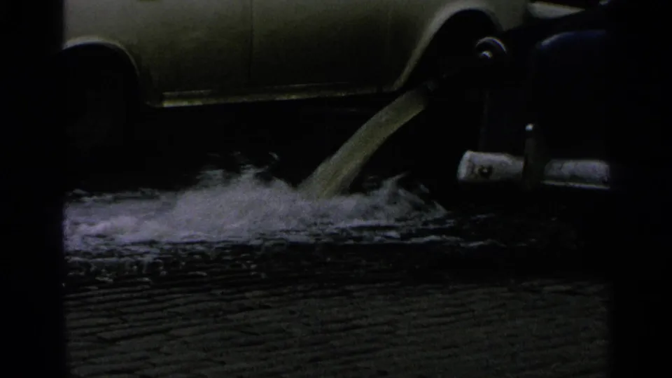 Foamy liquid pours onto wet pavement near the side of a parked car in 1965 Minnesota 8mm footage.