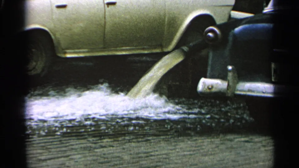 A stream of liquid splashes onto wet pavement beside a parked car, with dark film borders framing the scene.