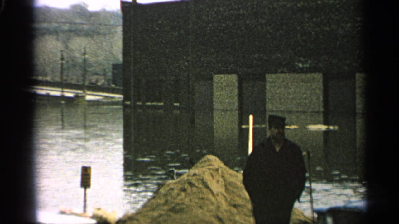 1965 Minnesota 8mm frame showing a man in dark outerwear near sandbags, floodwater, and a partially submerged waterfront structure.