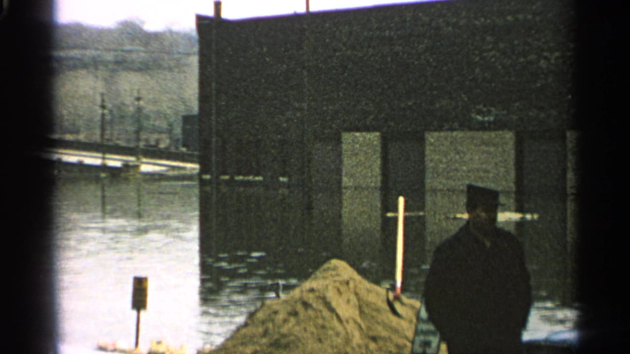 1965 Minnesota flood footage frame with a man beside sandbags, still high water, and dark structures along the flooded waterfront.