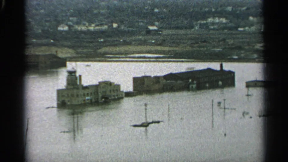 1965 Minnesota 8mm frame looking over floodwater toward partially surrounded waterfront buildings, a small boat, poles, and distant terrain.