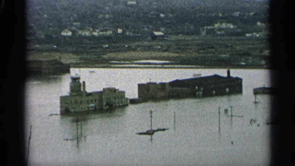 1965 Minnesota flood scene from an elevated home-movie viewpoint, showing inundated waterfront structures, a small boat, and distant hills.