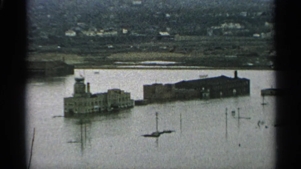 1965 Minnesota 8mm frame showing industrial waterfront buildings partly surrounded by floodwater, with distant shoreline structures behind them.