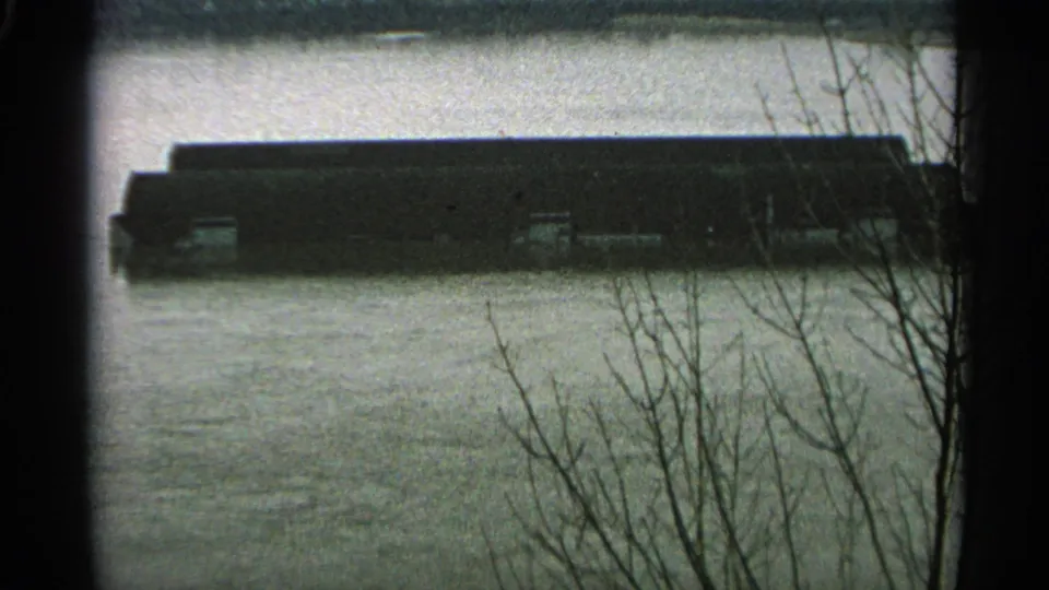 1965 Minnesota flood view of a long industrial structure standing in high water, seen through bare branches at the frame edge.