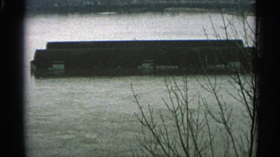 1965 Minnesota 8mm frame of floodwater around a low industrial building, with bare branches crossing the foreground.