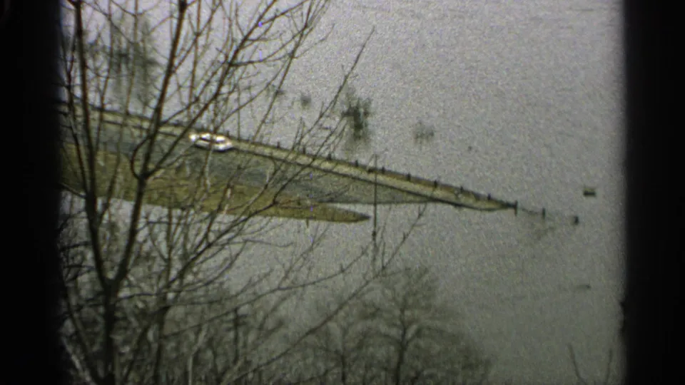 1965 Minnesota home-movie frame with bare trees, a submerged road, and a small vehicle beside floodwater.