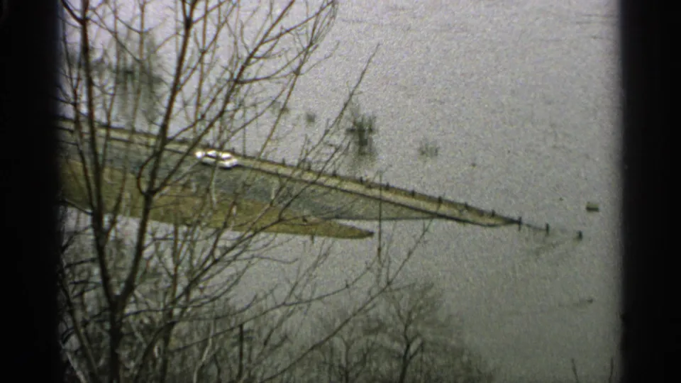Grainy 8mm view of a Minnesota flooded roadway, leafless trees in front, and a small vehicle near standing water.