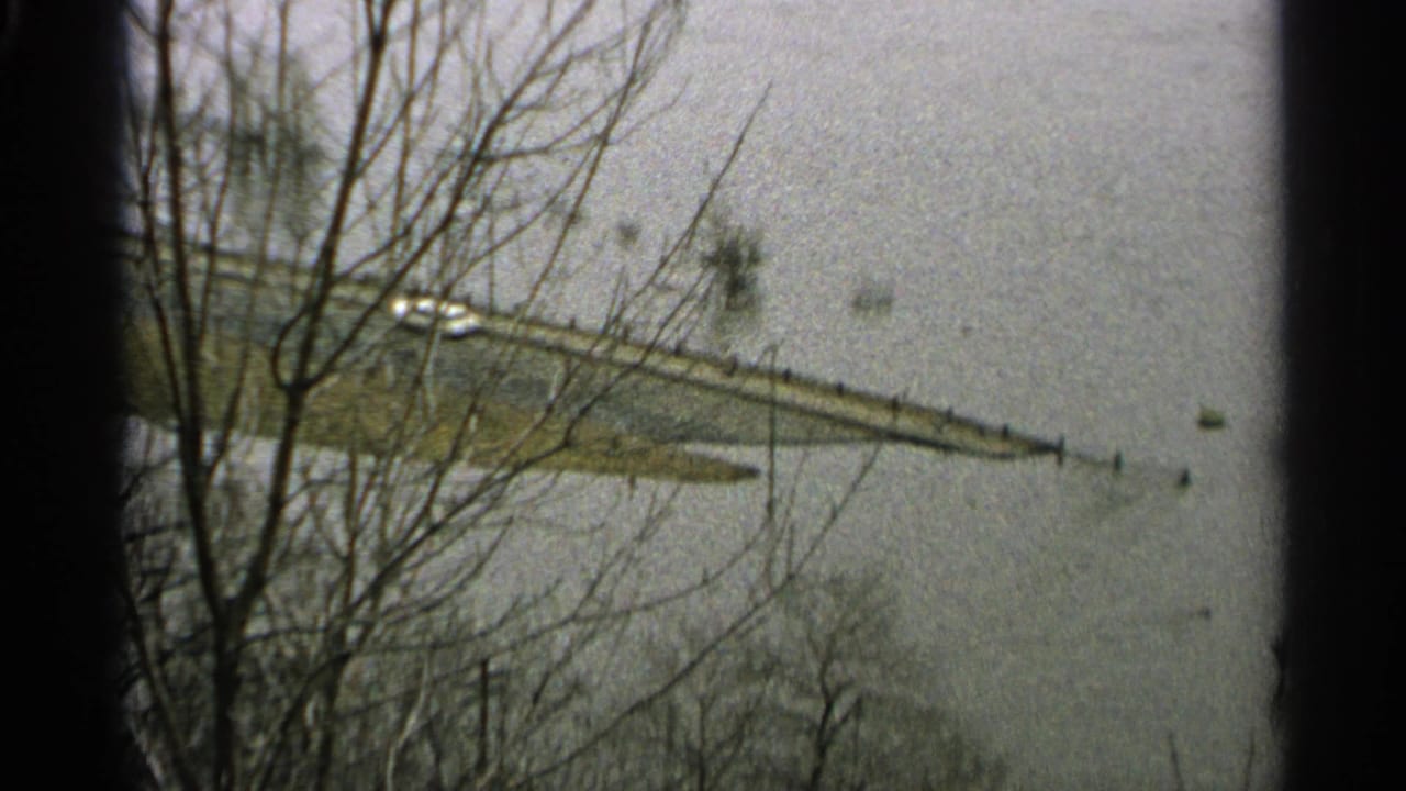 1965 Minnesota floodwater appears to cover low land around a narrow road or bridge, with a white car visible near leafless trees.