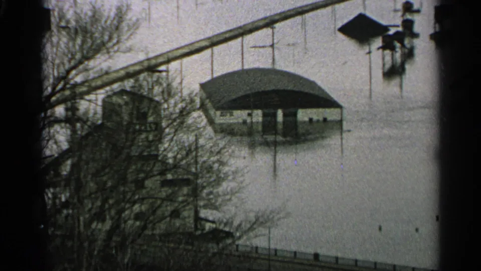 Floodwater surrounds a low curved-roof building in the Minnesota scene, with bare trees, a long bridge or walkway, and nearby posts visible.