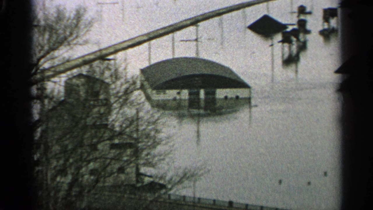 1965 Minnesota 8mm frame showing murky floodwater around low buildings, bare trees, posts, and an elevated crossing in the distance.