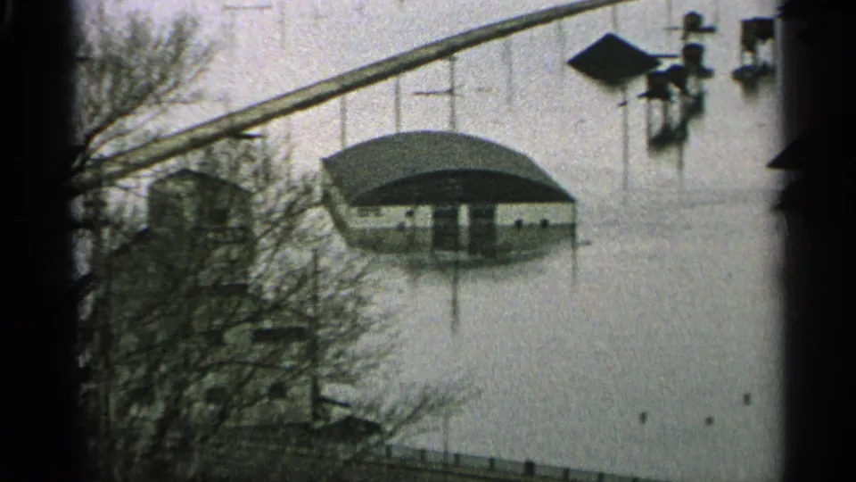 1965 Minnesota 8mm frame showing murky floodwater around low buildings, bare trees, posts, and an elevated crossing in the distance.