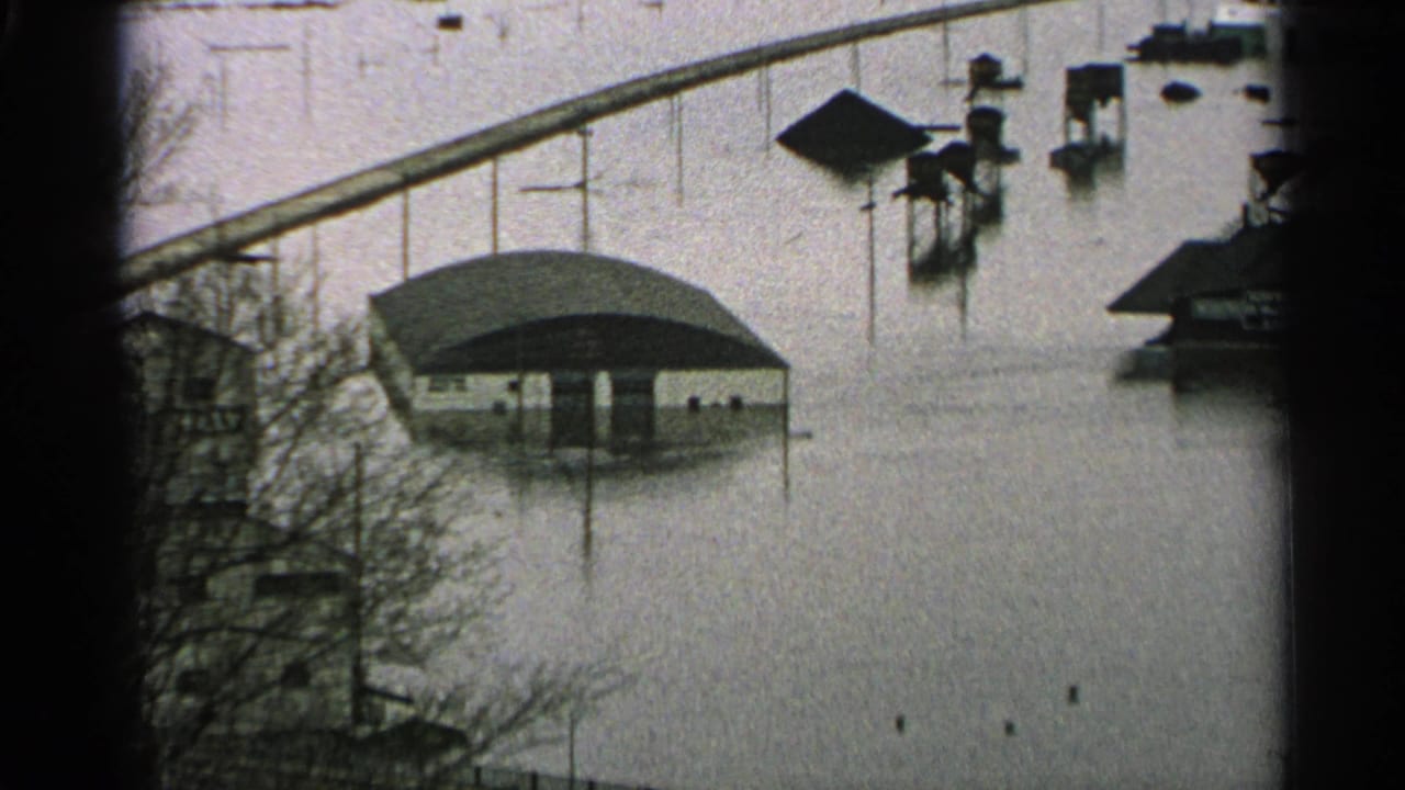 1965 Minnesota flood scene with a low roofed structure, posts, and nearby buildings surrounded by high murky water.