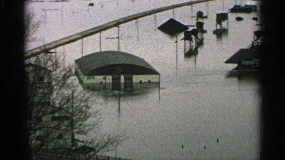 1965 Minnesota flood scene with a low roofed structure, posts, and nearby buildings surrounded by high murky water.