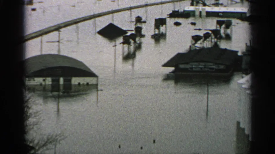 1965 Minnesota 8mm view of floodwater surrounding multiple roofed structures, with posts and bare branches visible in the foreground.