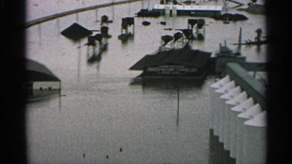 1965 Minnesota flood footage shows a dark-roofed building, surrounding posts, and high water extending across the scene.
