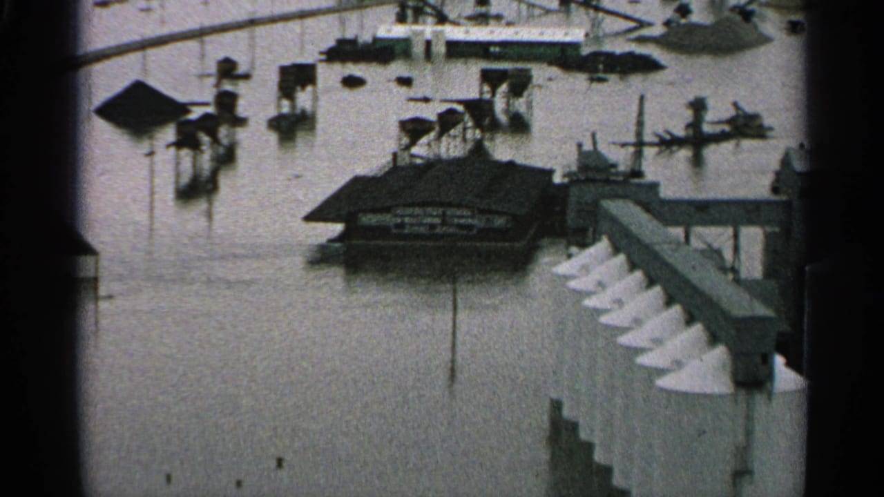 1965 Minnesota 8mm frame showing floodwater around an industrial building, elevated walkways, pilings, and white conical silo tops.