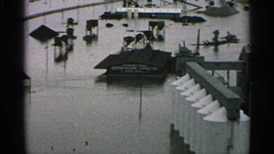 1965 Minnesota 8mm frame showing floodwater around an industrial building, elevated walkways, pilings, and white conical silo tops.