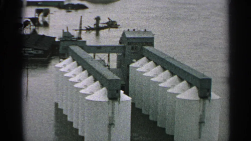 1965 Minnesota 8mm frame showing rows of white conical silo tops beside floodwater, with industrial buildings and dock-like structures behind them.