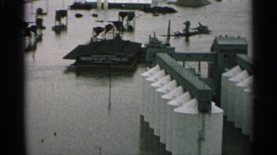 1965 Minnesota frame showing floodwater across an industrial site, with a low building, elevated walkways, and white cone-topped silos visible.