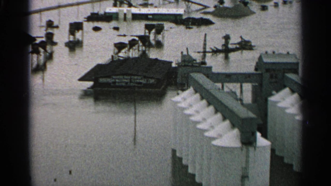 1965 Minnesota flood scene showing an industrial waterfront, partly submerged boats, dock structures, and murky water around riverfront buildings.