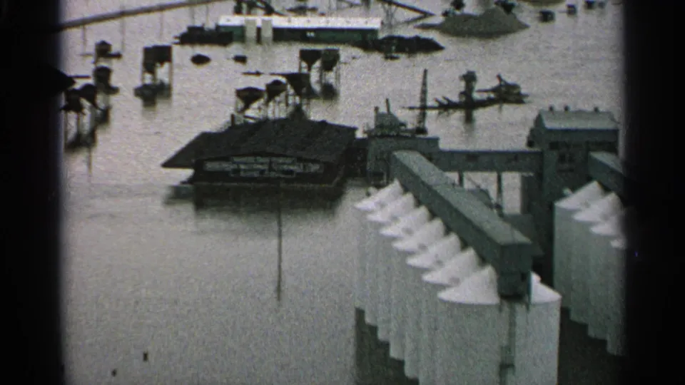 1965 Minnesota flood scene showing an industrial waterfront, partly submerged boats, dock structures, and murky water around riverfront buildings.