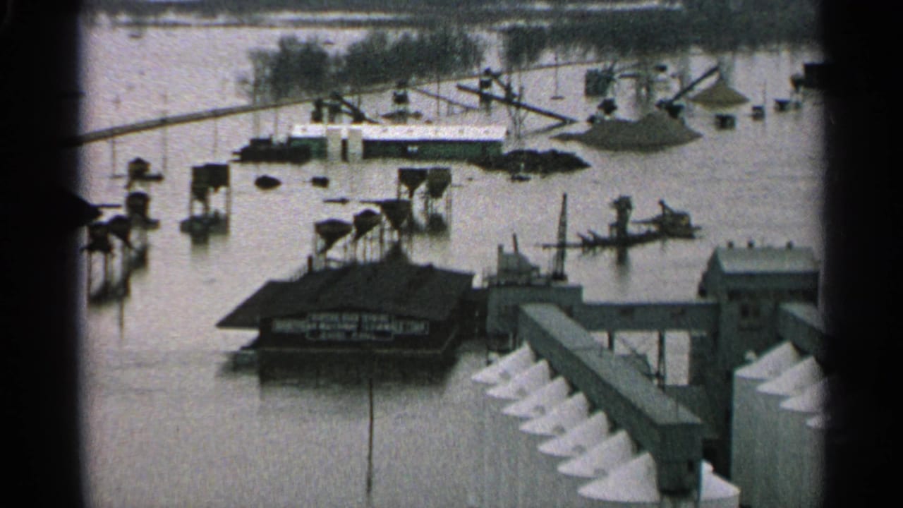 Floodwater covers docks and surrounds boats near industrial waterfront buildings.