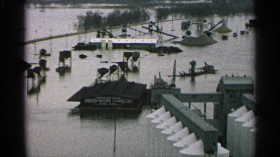 1965 Minnesota riverfront flooding with boats, dock pilings, a low industrial building, and a conveyor-like structure partly surrounded by water.
