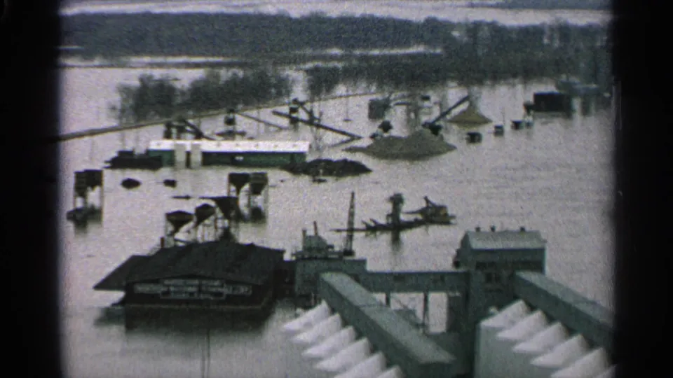 1965 Minnesota elevated view of floodwater across an industrial waterfront, with boats, dock remnants, a roofed structure, and conveyor equipment visible.