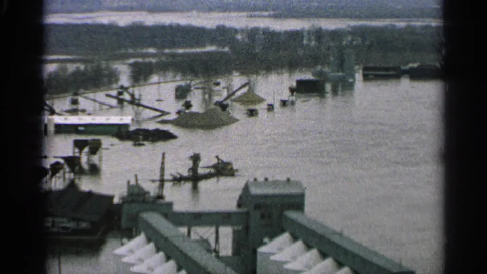 1965 Minnesota wide view of a flooded industrial waterfront, with boats and dock structures partly submerged and distant trees beyond the water.