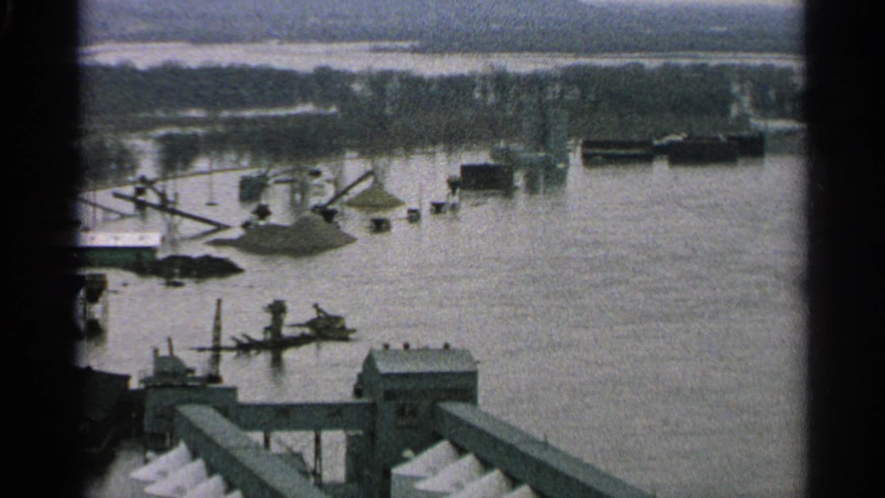 1965 Minnesota 8mm frame showing a flooded industrial waterfront with docks, cranes, barges, low buildings, and a hazy far shoreline.