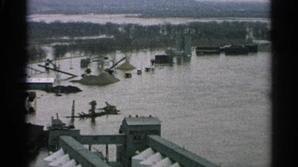 1965 Minnesota 8mm frame showing a flooded industrial waterfront with docks, cranes, barges, low buildings, and a hazy far shoreline.