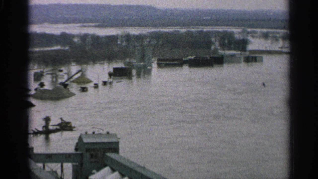Elevated 1965 Minnesota view of a flooded port or shipyard area with docked vessels, cranes, industrial sheds, trees, and distant shoreline.