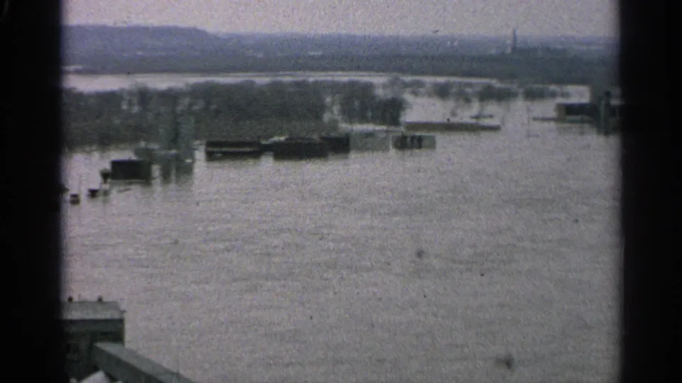 1965 Minnesota 8mm frame with floodwater across a port landscape, showing barges, industrial buildings, bare trees, and a faint skyline beyond.