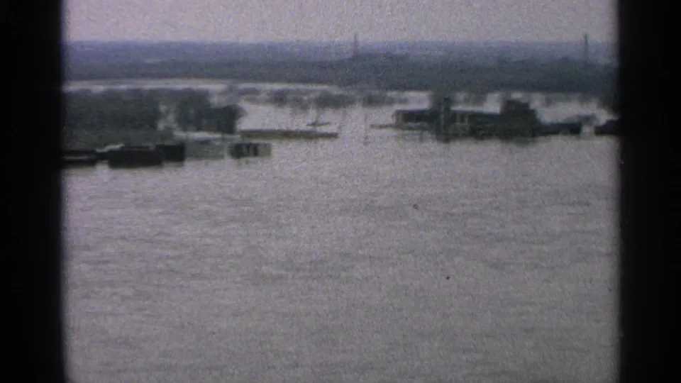 Soft 1965 Minnesota frame showing a broad flooded industrial waterfront with barges, low dock structures, distant trees, and a hazy skyline.