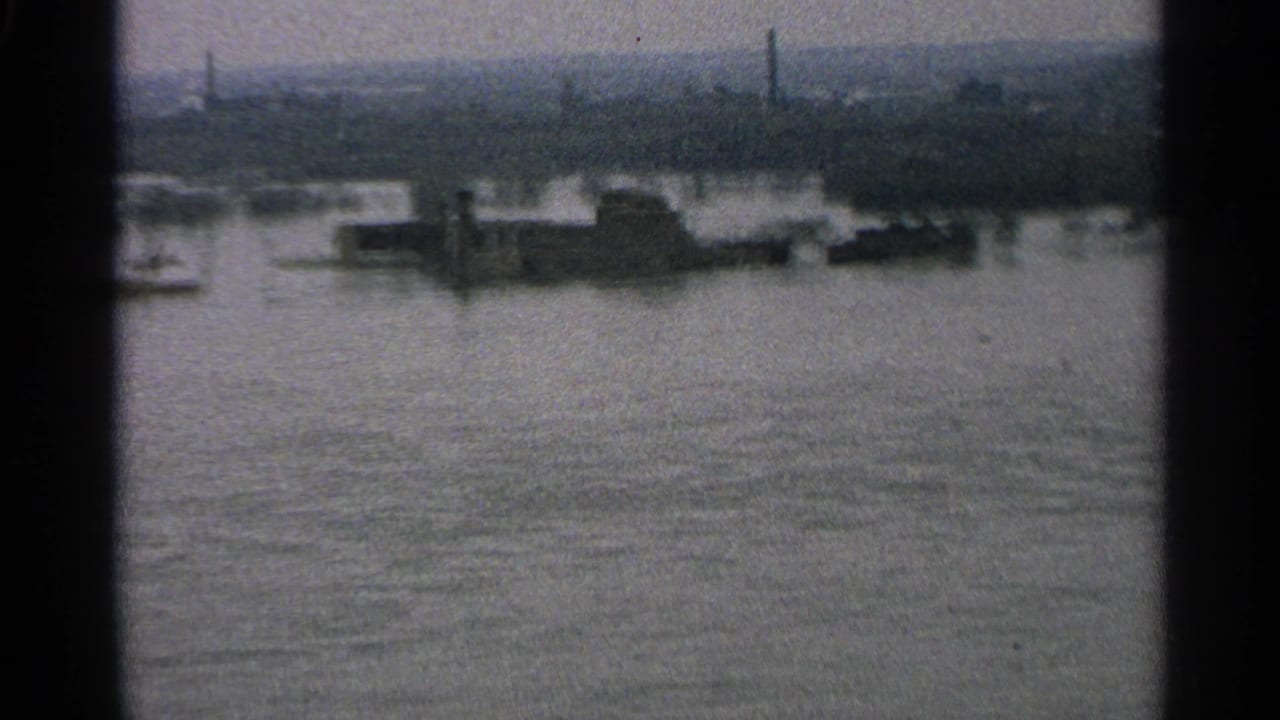 1965 Minnesota 8mm frame showing floodwater across a shoreline with low structures and smokestacks in the distance.
