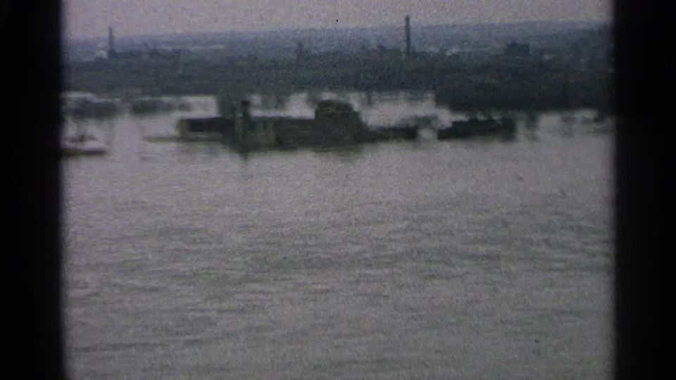 1965 Minnesota 8mm frame showing floodwater across a shoreline with low structures and smokestacks in the distance.