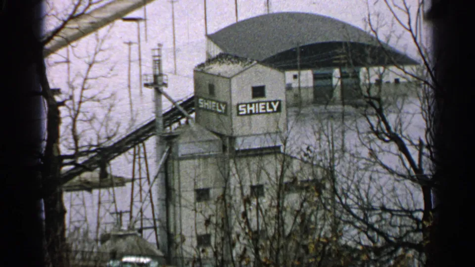 1965 Minnesota ski-area frame showing a building marked SHIELY, lift equipment, bare trees, and winter ground.