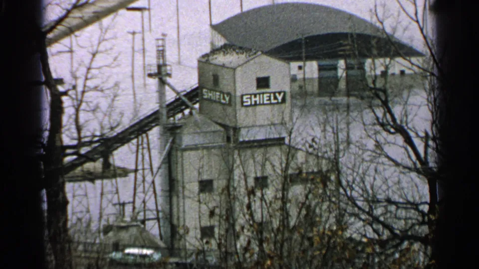 1965 Minnesota 8mm frame of a ski-area structure with SHIELY signage, lift cables, poles, and leafless trees.