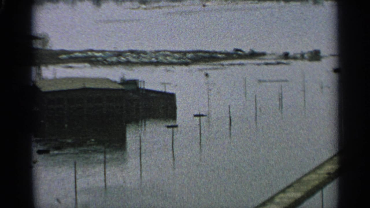 1965 Minnesota 8mm frame showing a flooded waterway beside a low waterfront building, with utility poles protruding from the water.