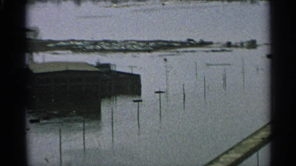 1965 Minnesota 8mm frame showing a flooded waterway beside a low waterfront building, with utility poles protruding from the water.