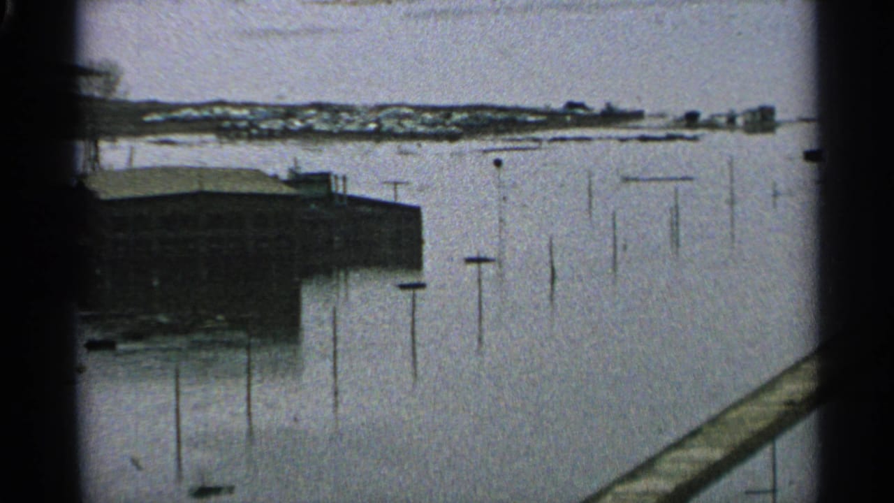 1965 Minnesota flood view with a shoreline building, utility poles in high water, and a diagonal railing or dock in the foreground.