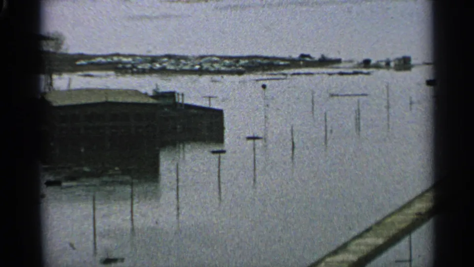 1965 Minnesota flood view with a shoreline building, utility poles in high water, and a diagonal railing or dock in the foreground.