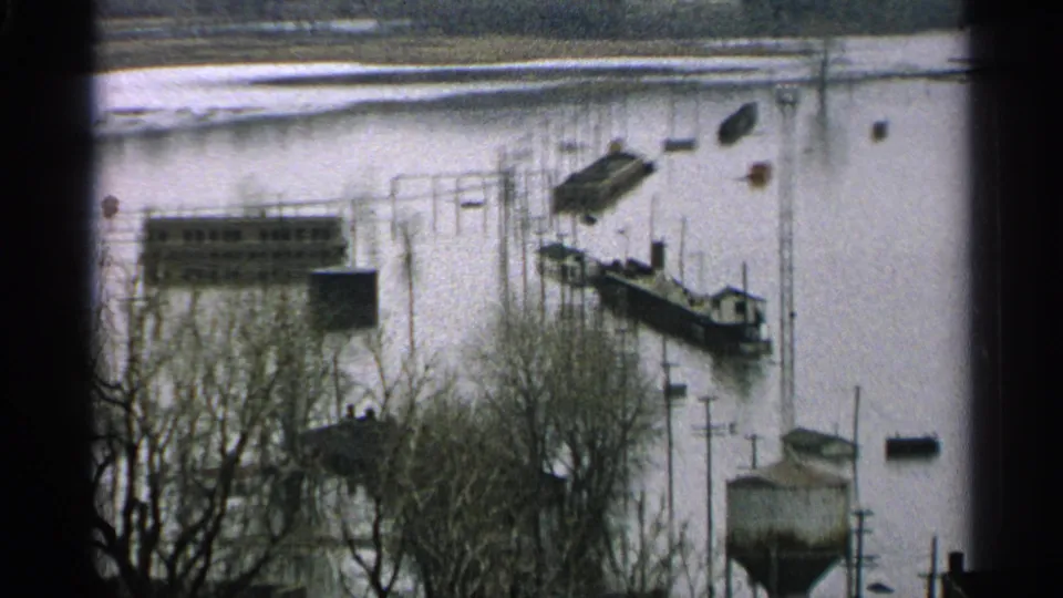 1965 Minnesota flood scene with boats near submerged docks, leafless trees in the foreground, and utility poles standing in high water.