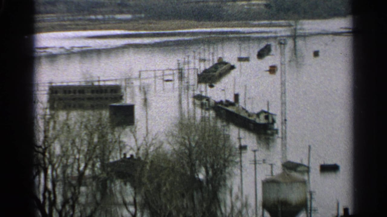 Minnesota 1965 8mm frame showing floodwater around docks, boats, bare trees, and low waterfront buildings.