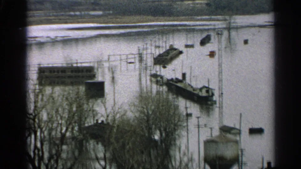 Minnesota 1965 8mm frame showing floodwater around docks, boats, bare trees, and low waterfront buildings.