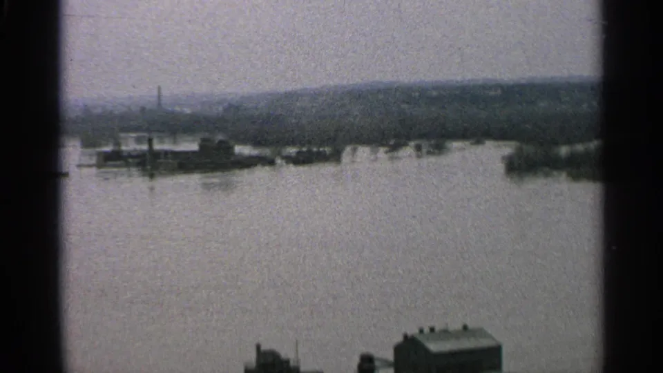 Wide 8mm view of a Minnesota flood scene with high water across a riverfront and distant hills beyond.