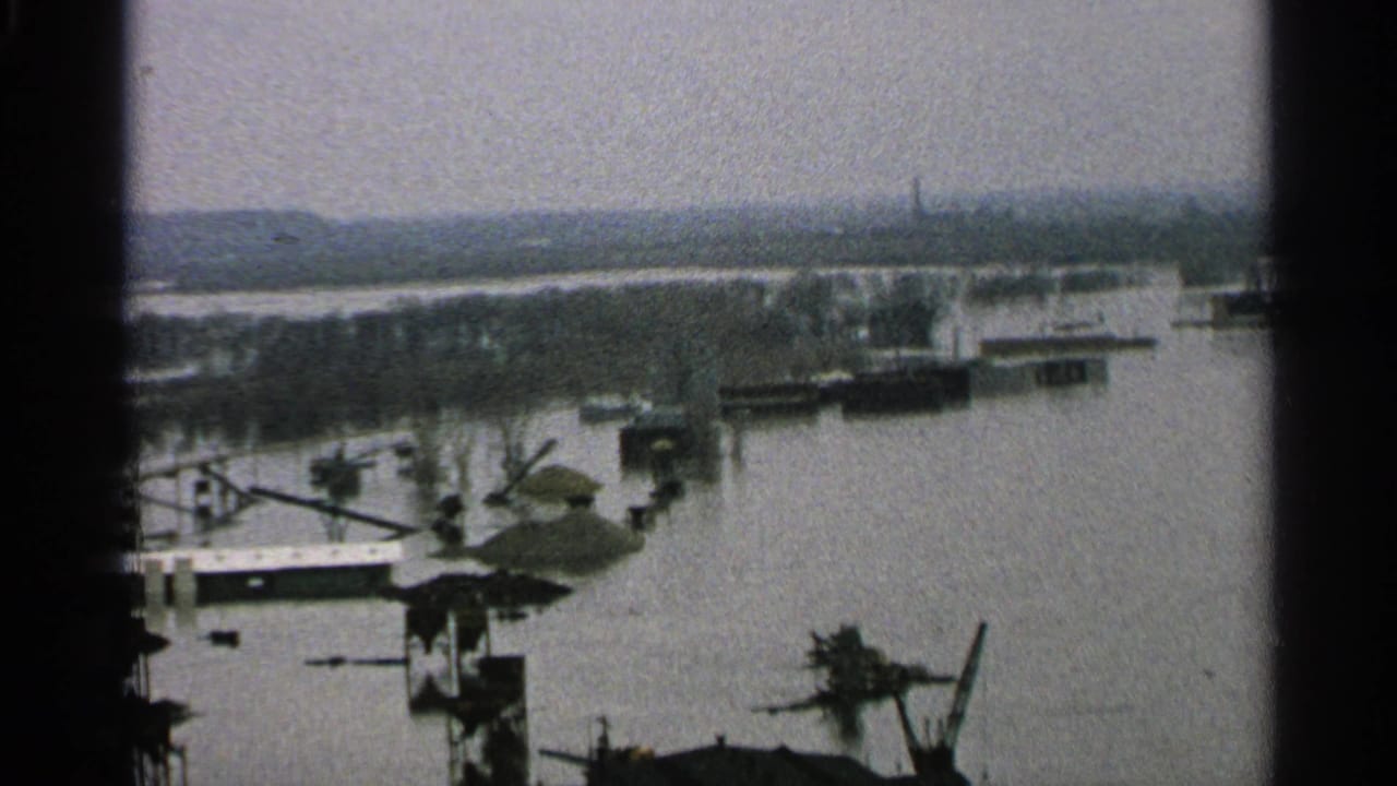 1965 Minnesota 8mm frame showing a flooded industrial edge with docks, low structures, crane-like equipment, and a hazy shoreline.