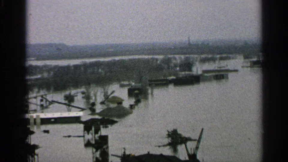 1965 Minnesota 8mm frame showing a flooded industrial edge with docks, low structures, crane-like equipment, and a hazy shoreline.